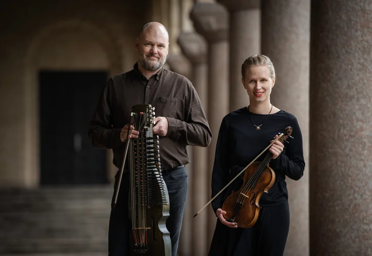 En man och en kvinna står sida vid sida med stråkinstrument i en pelargång.