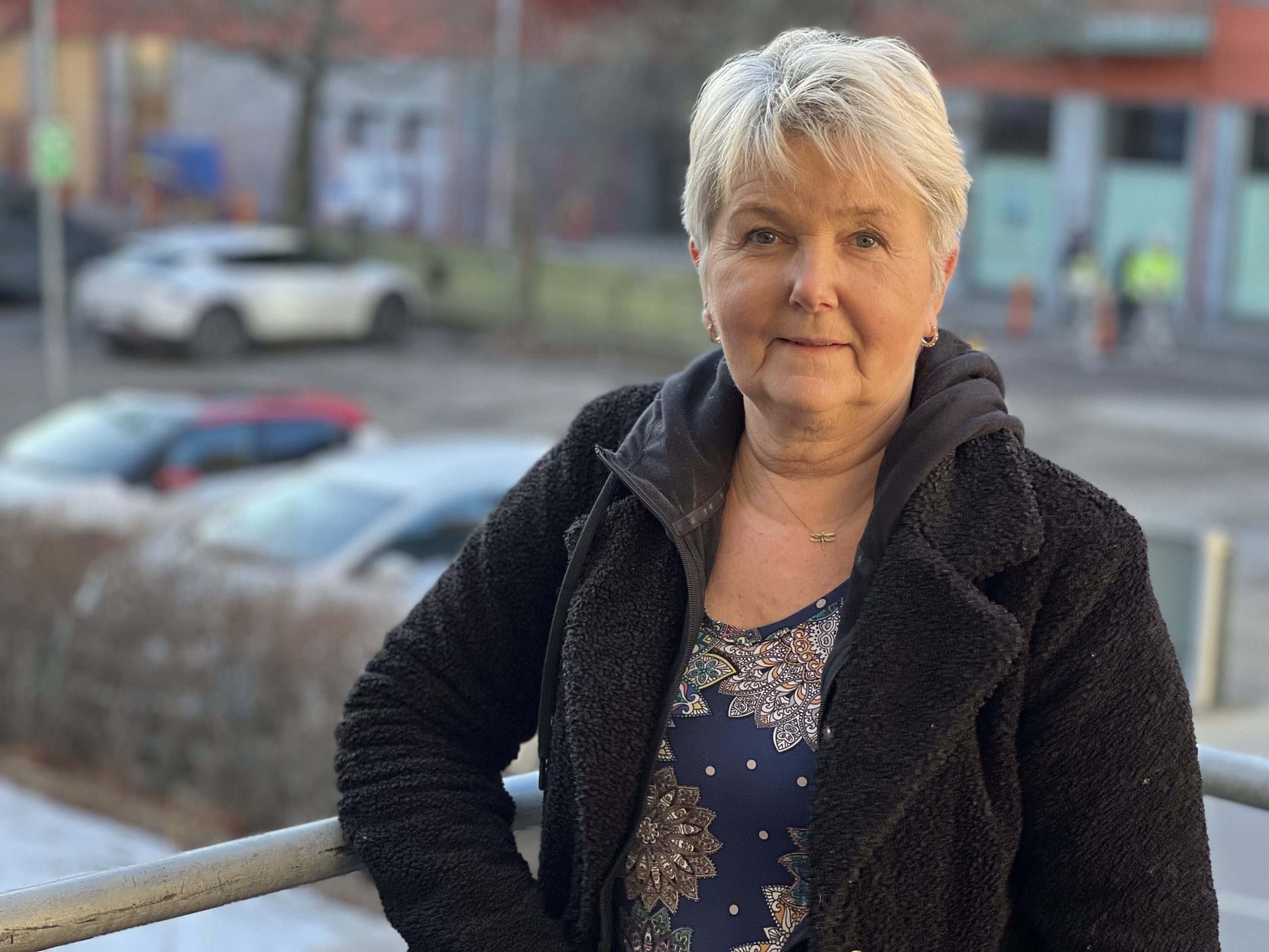 En äldre kvinna med kort grått hår står utomhus vid ett räcke, med parkerade bilar och byggnader bakom sig.