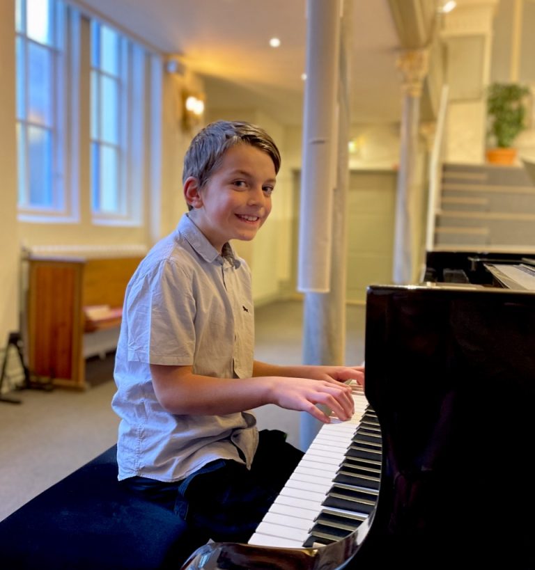 En pojke i ljus skjorta ler medan han spelar på en flygel i ett ljust rum med stora fönster.