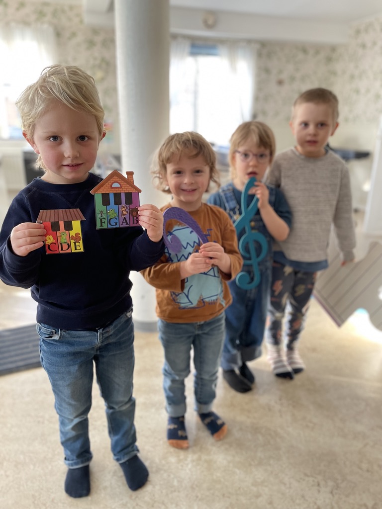 Fyra små barn står inomhus och håller i färgglad rekvisita med musiktema och ler mot kameran.