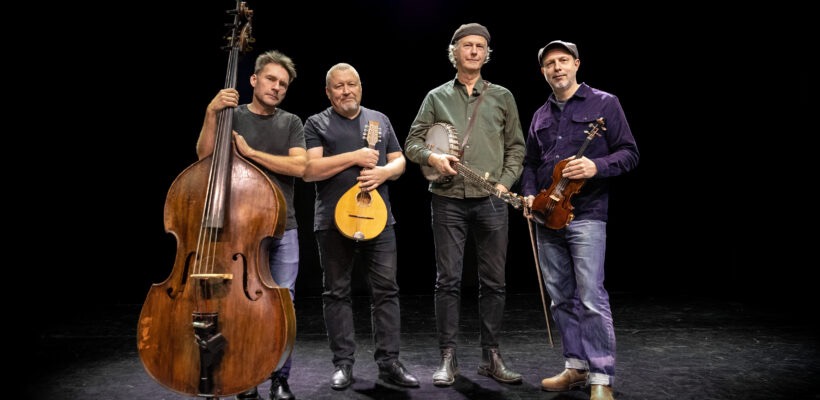 Fyra män står tillsammans och håller i en kontrabas, mandolin, banjo och violin mot en svart bakgrund.
