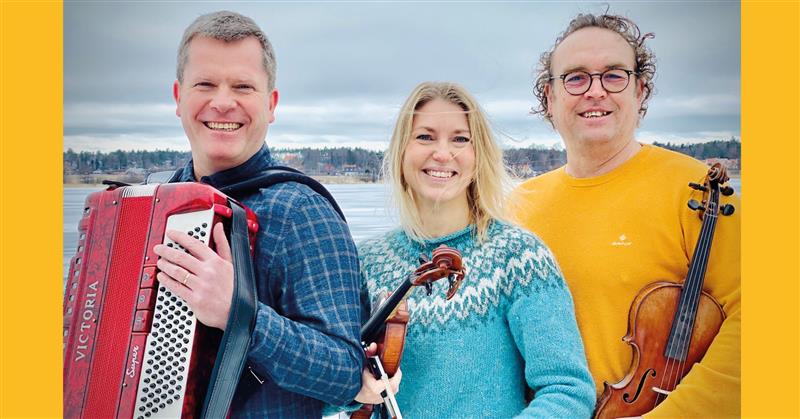 Tre personer ler utomhus med musikinstrument i händerna: ett dragspel och två violiner, med en sjö i bakgrunden.