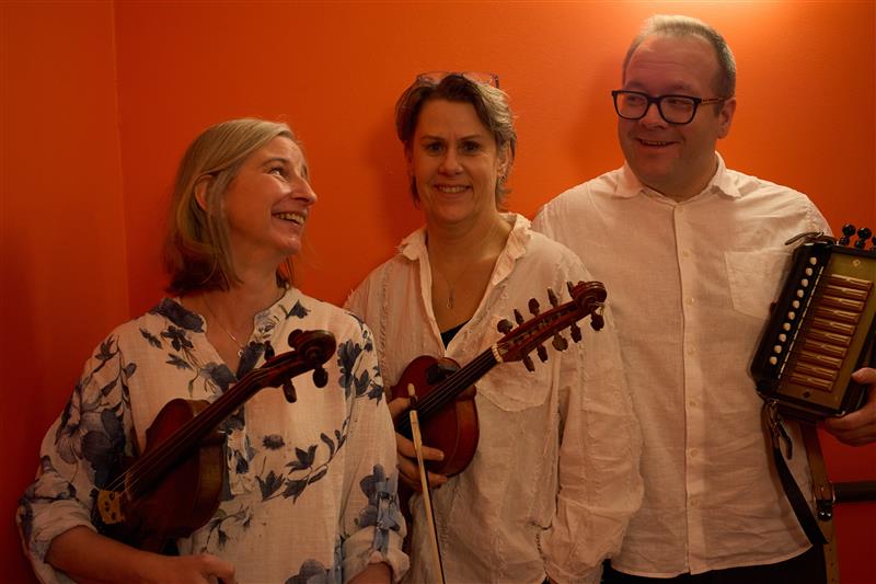 Tre personer i vita skjortor håller i musikinstrument och ler mot en orange vägg.