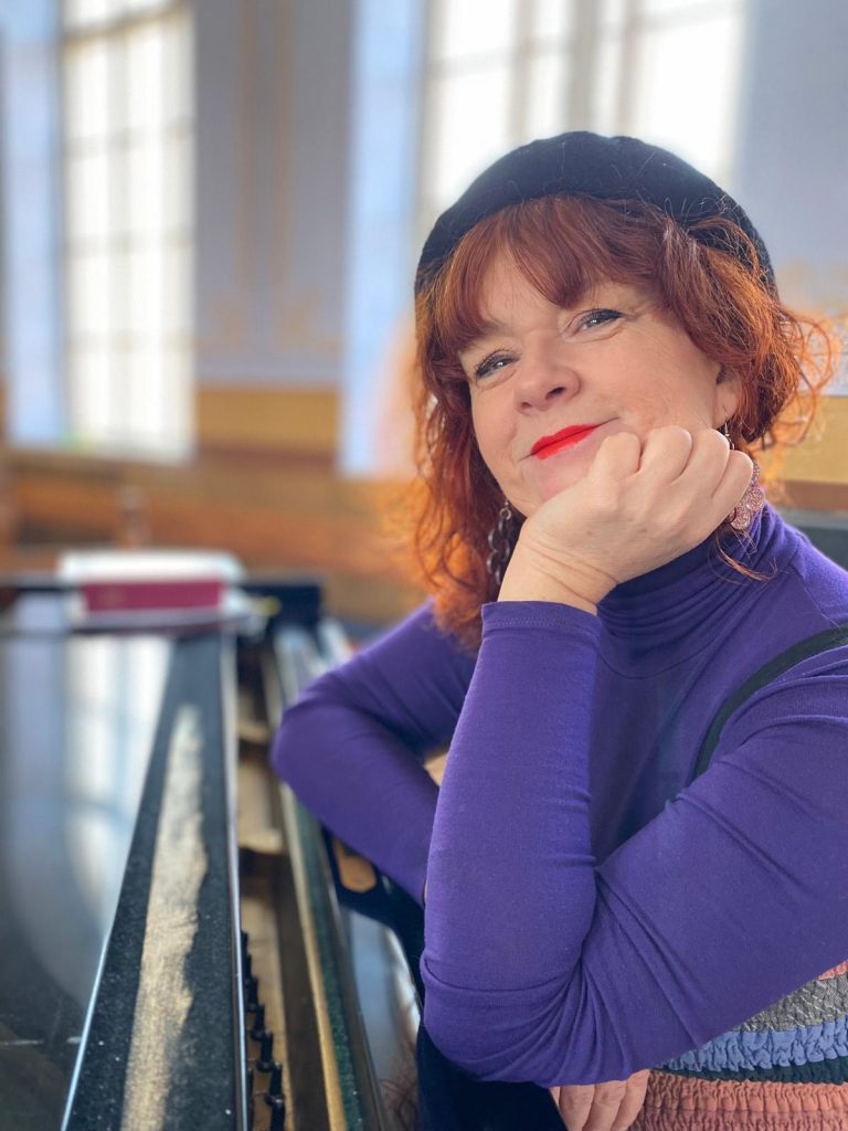 En kvinna med rött hår och svart basker lutar sig mot ett piano och ler eftertänksamt i ett solbelyst rum.