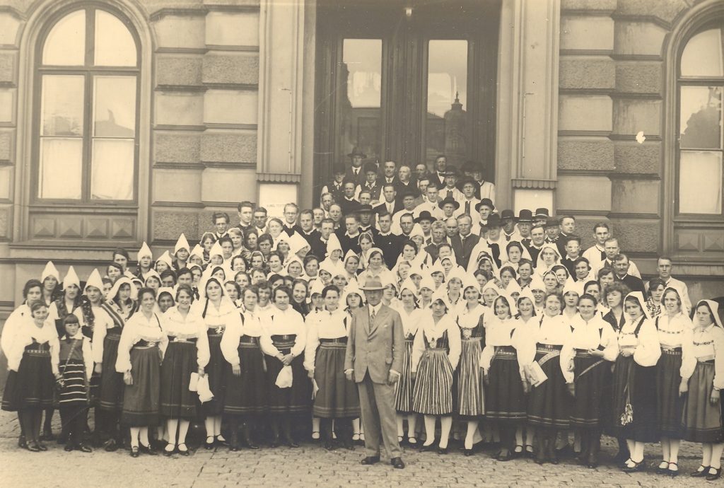 En stor grupp i traditionella kläder poserar på en trappa utanför en historisk byggnad för ett gammalt gruppfoto.