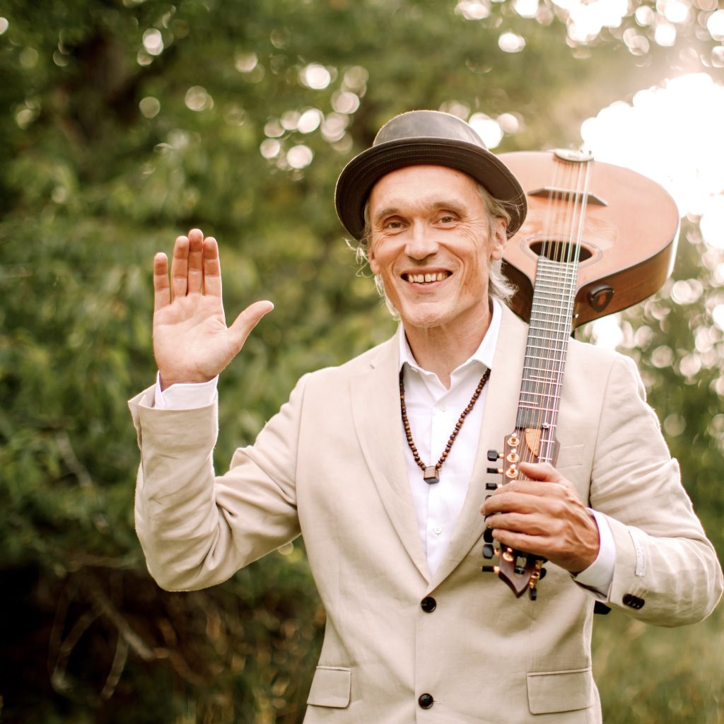 Leende man i beige kostym med gitarr på axeln vinkar utomhus i naturligt ljus.