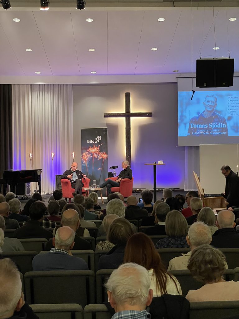 Två personer talar på scenen framför en publik, med ett stort kors och en skärm bakom sig.