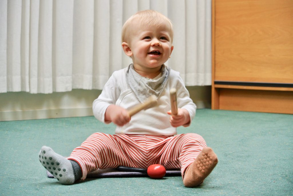 Leende bebis sitter på matta med trumpinnar i handen och ett rött leksaksägg i närheten.