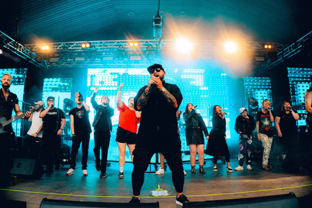 En man sjunger på scenen med en grupp människor bakom sig, i starkt konsertljus.