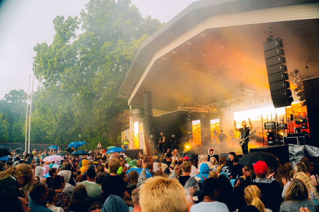 Publik som tittar på en livlig utomhuskonsert på en scen med färgglada lampor och träd i bakgrunden.