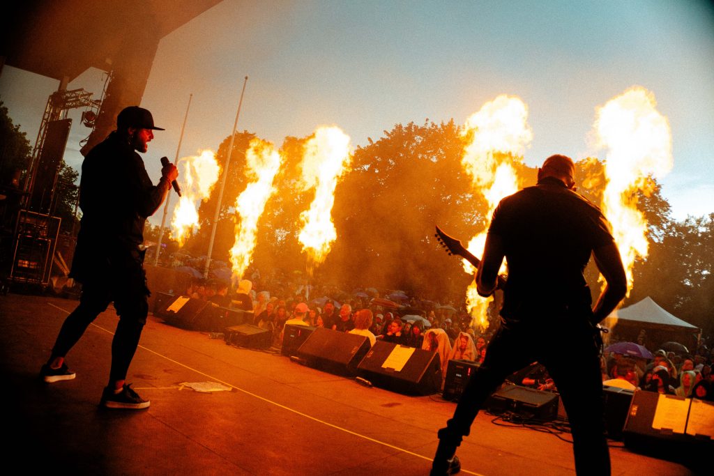 Två musiker uppträder på scen med stora eldslågor bakom sig inför en publik i solnedgången.