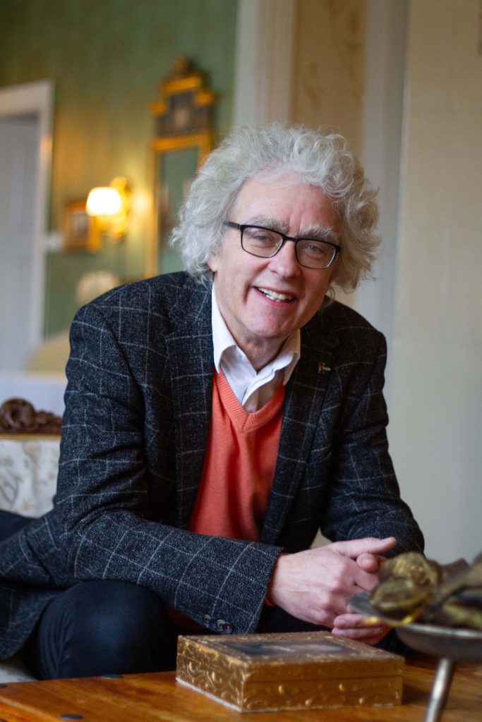 En äldre man med lockigt grått hår och glasögon ler när han sitter inomhus, klädd i kavaj och orange tröja.