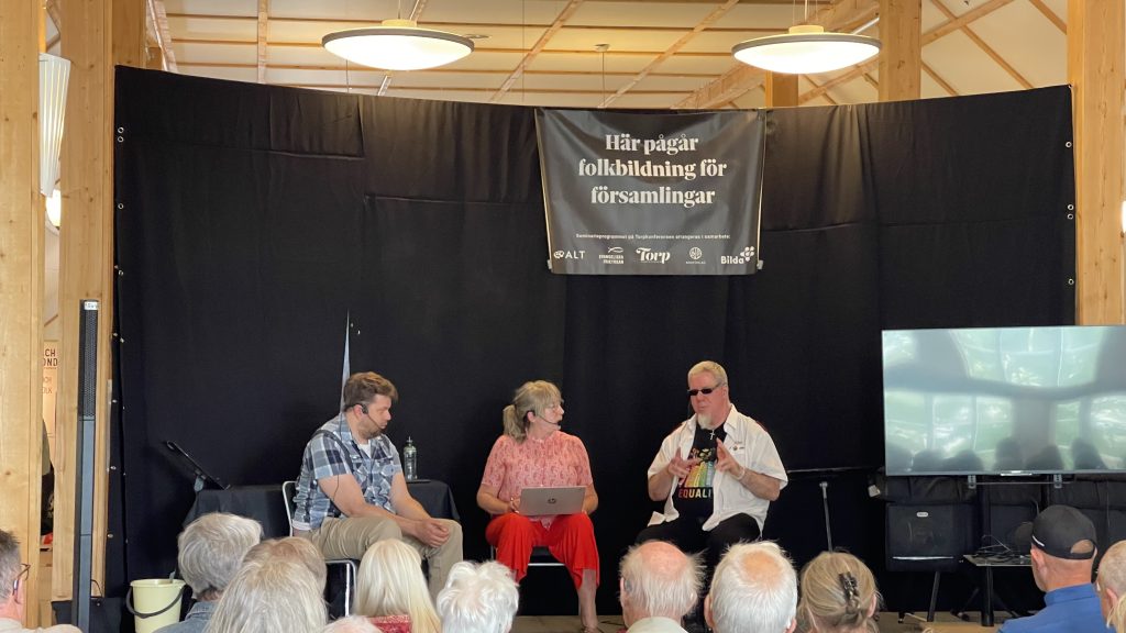 Tre personer sitter på scenen och diskuterar inför publik, med en svart banderoll hängande bakom sig.