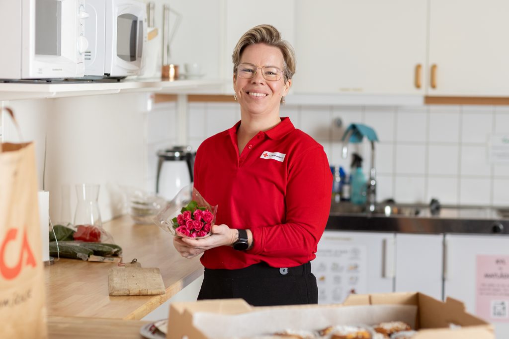 Leende person i röd skjorta håller blommor i ett kök med bakverk på disken.