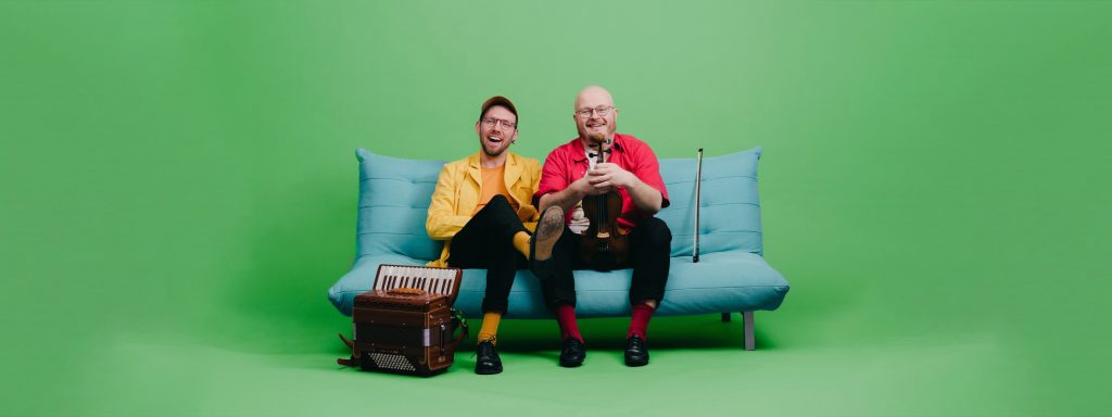 Två leende män sitter i en blå soffa mot en grön bakgrund, håller i instrument och tittar in i kameran.