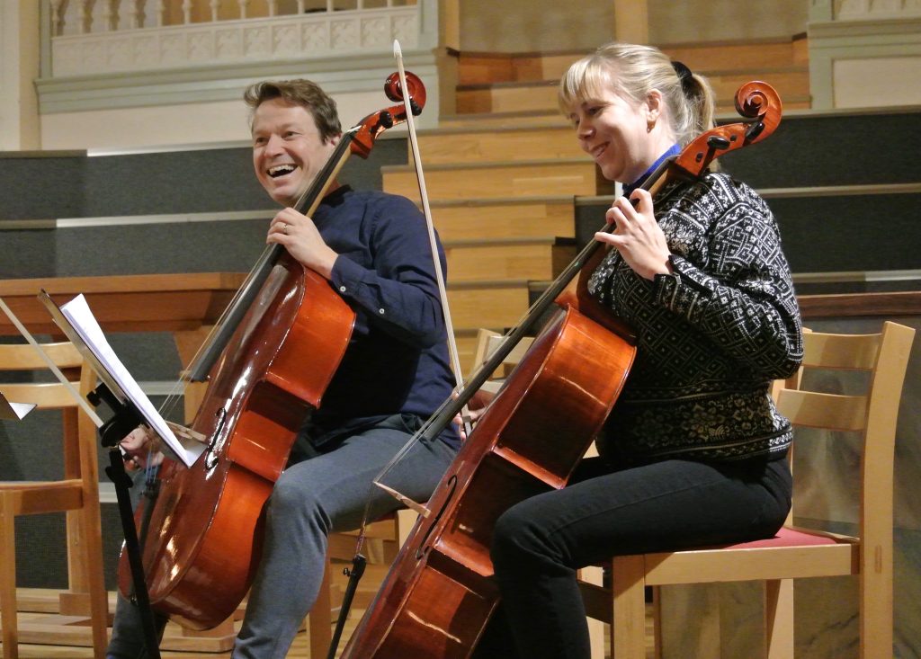 Två personer sitter på stolar, ler och spelar cello i ett varmt upplyst rum med träaccenter.