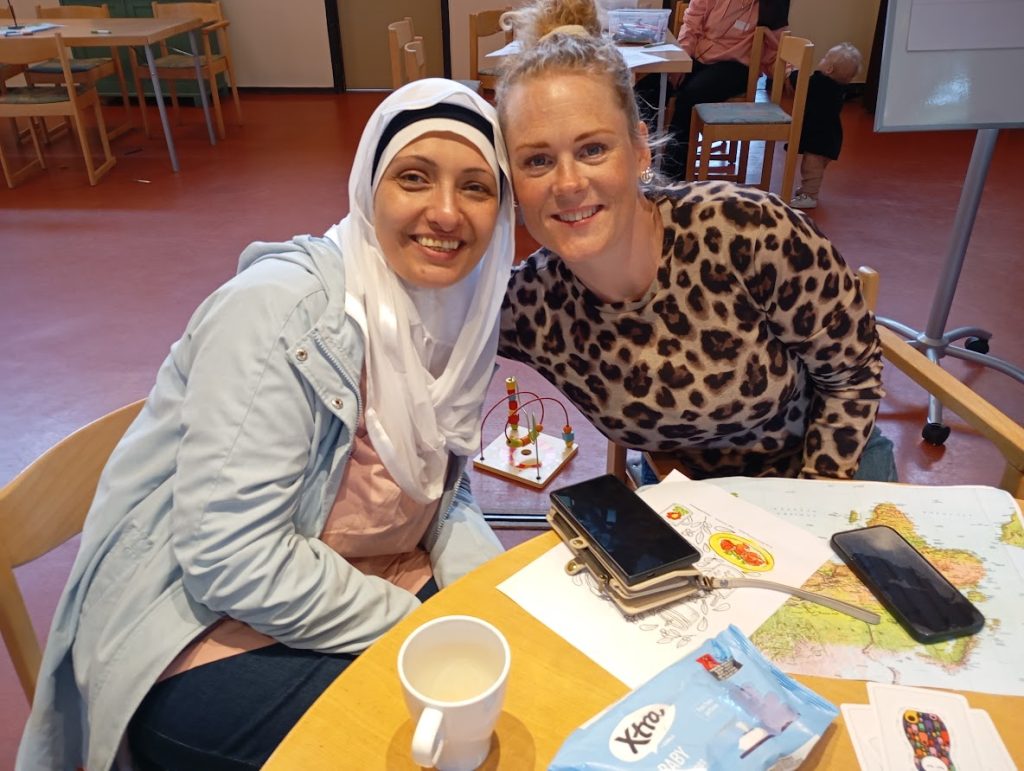 Två kvinnor ler och sitter tillsammans vid ett bord med papper, en kopp och telefoner i ett ljust rum.