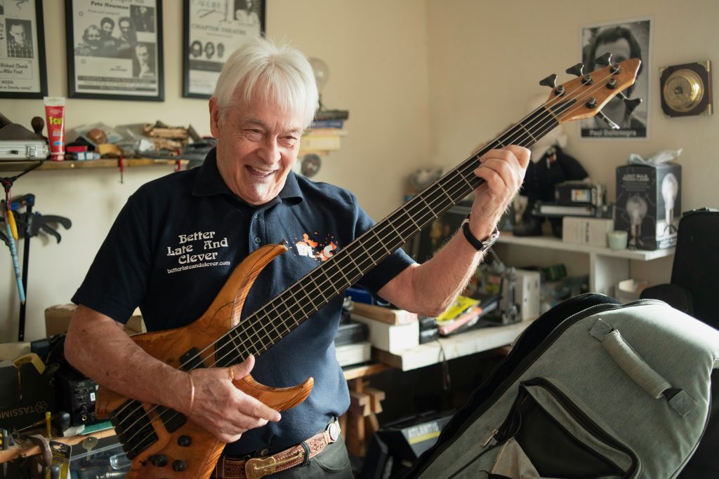 En leende äldre man spelar på en elektrisk basgitarr i trä i ett rum fyllt av musikminnen och böcker.