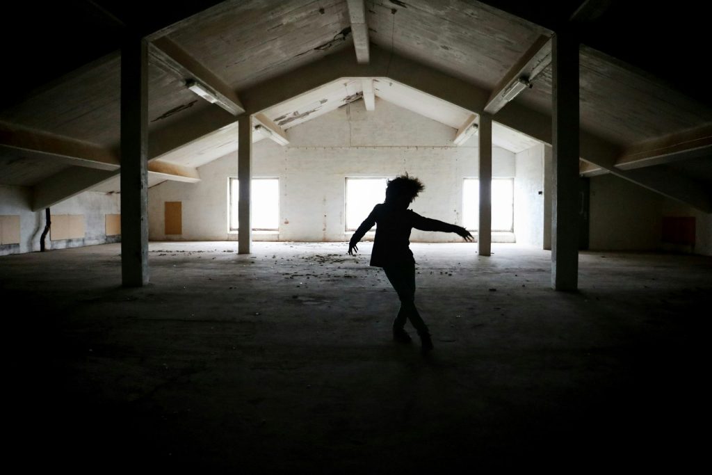 Silhuett av en person som dansar på en tom, svagt upplyst vind med solljus som strömmar in genom fönstren.