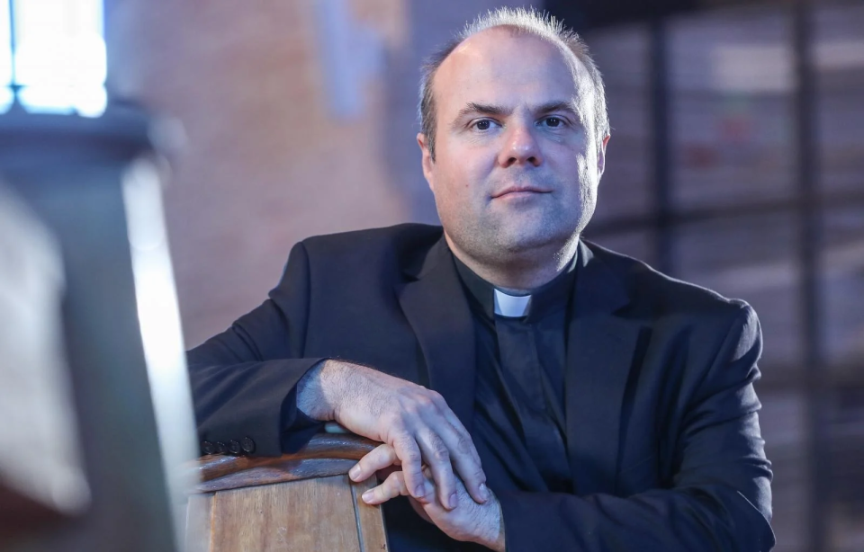 En präst i svart kostym sitter i en kyrka, vilar armen på en träbänk och tittar rakt fram.