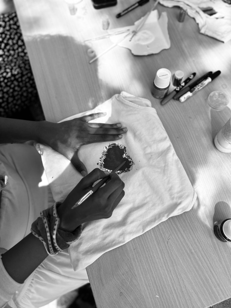 En person dekorerar en vit t-shirt med ett hjärtmotiv med hjälp av tuschpennor och färg vid ett bord.