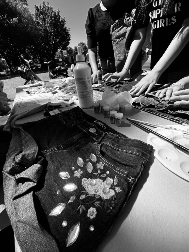 En grupp dekorerar jeansshorts med färg och ädelstenar utomhus en solig dag.