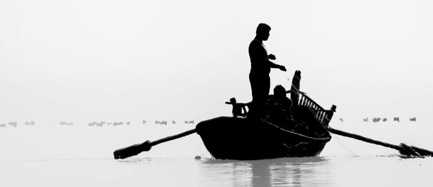 Silhuett av en person som står på en roddbåt av trä i lugnt vatten och håller i ett fiskenät.