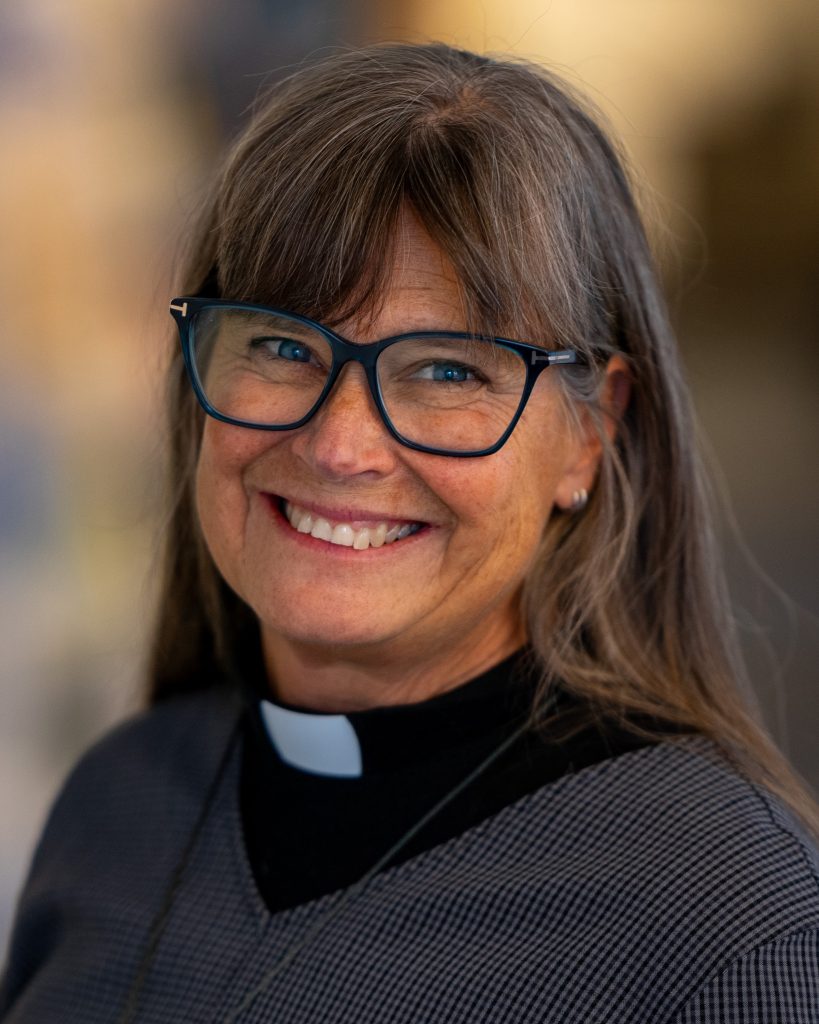 Leende kvinna med glasögon och grått hår, klädd i prästkrage och mörk tröja, inomhus.