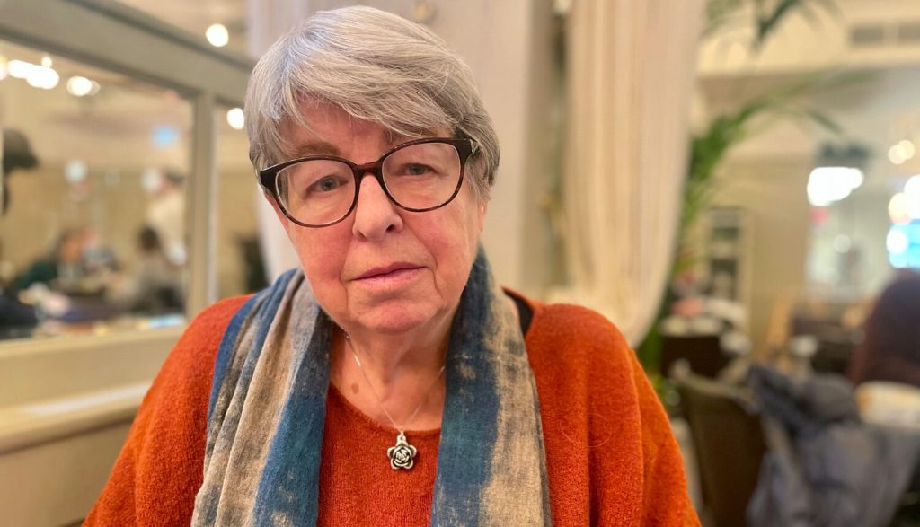 En äldre kvinna med grått hår, glasögon och halsduk sitter inomhus och tittar in i kameran.