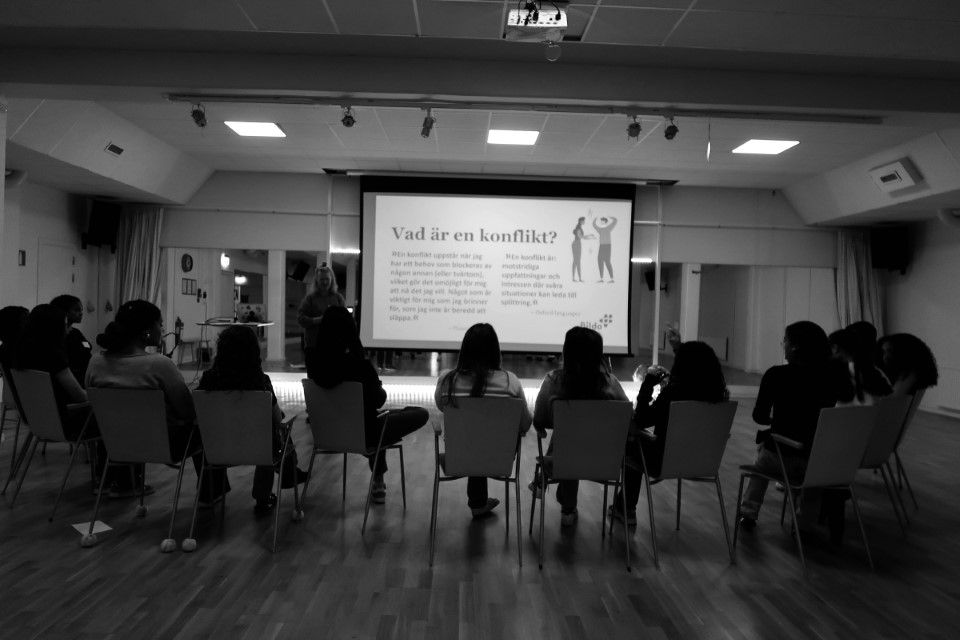 En grupp människor sitter framför en presentationsskärm i ett modernt och rymligt rum.