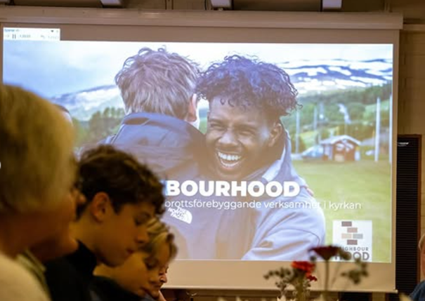 En projicerad bild visar en leende person som kramar ett barn med ordet "NEIGHBOURHOOD" på skärmen.