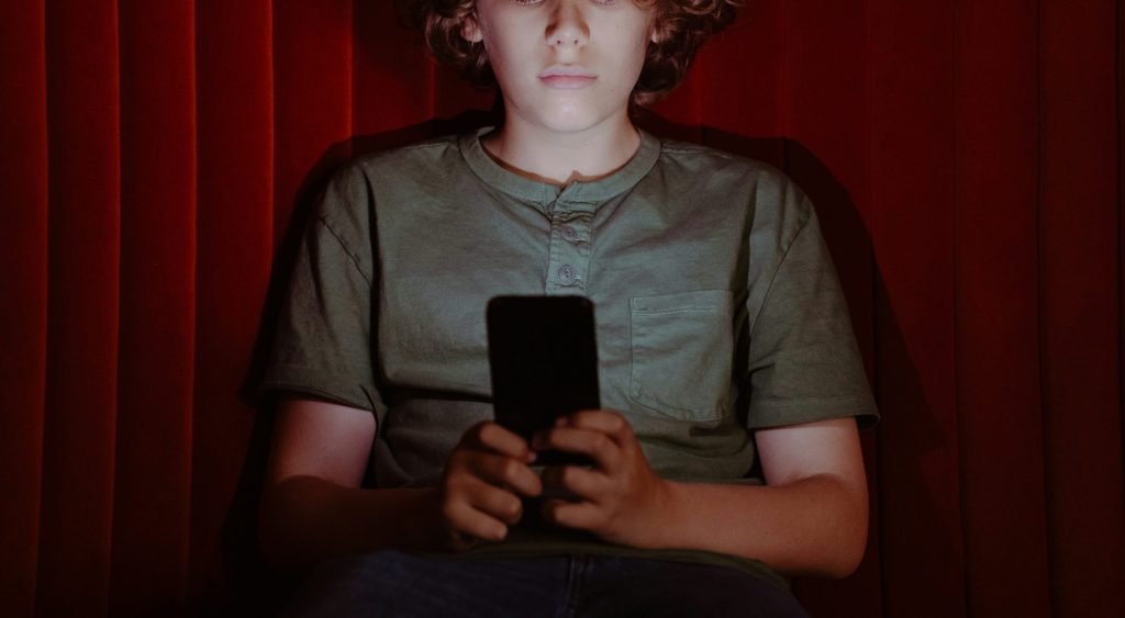 En pojke med lockigt hår sitter mot en röd gardin och tittar på en smartphone i sina händer.