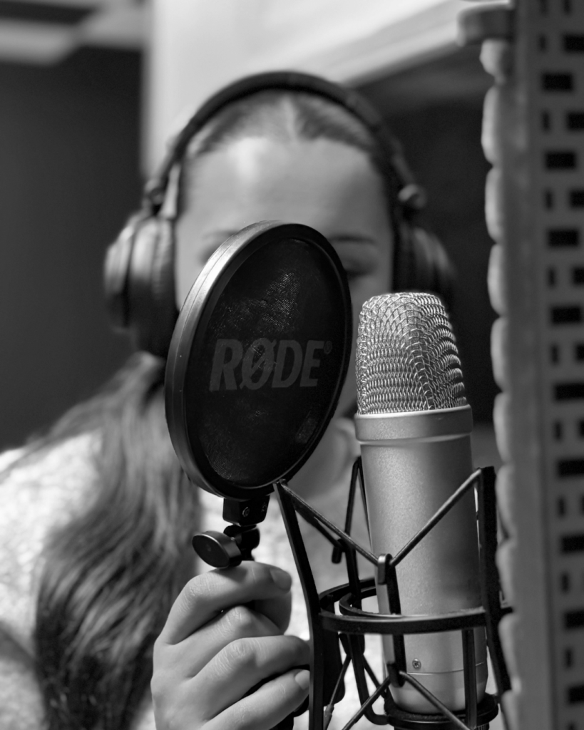 Person med hörlurar som sjunger i en mikrofon bakom ett popfilter i en inspelningsstudio, svartvitt.