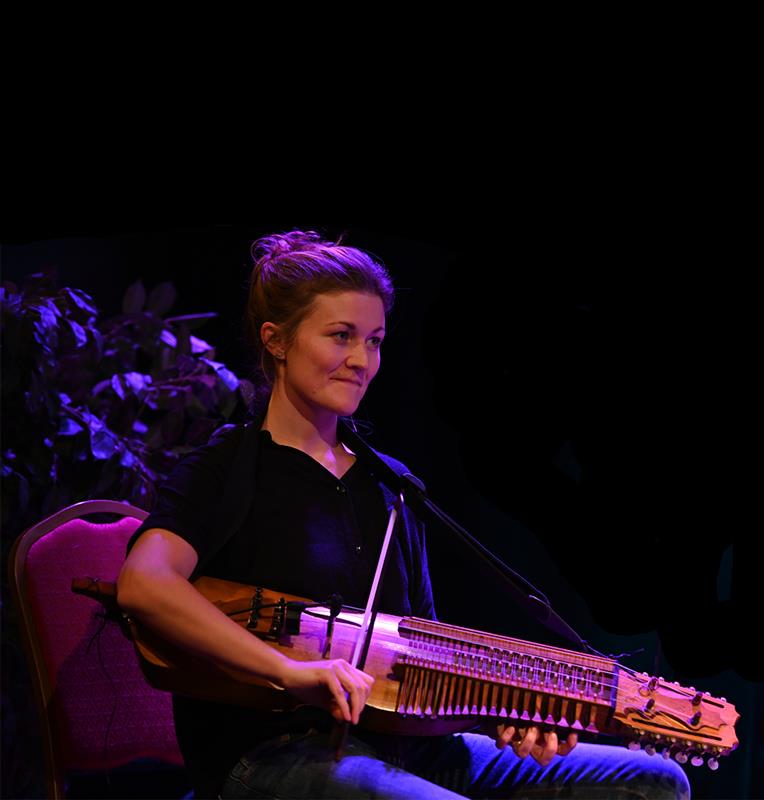En kvinna sitter och spelar nyckelharpa med stråke mot en mörk bakgrund.