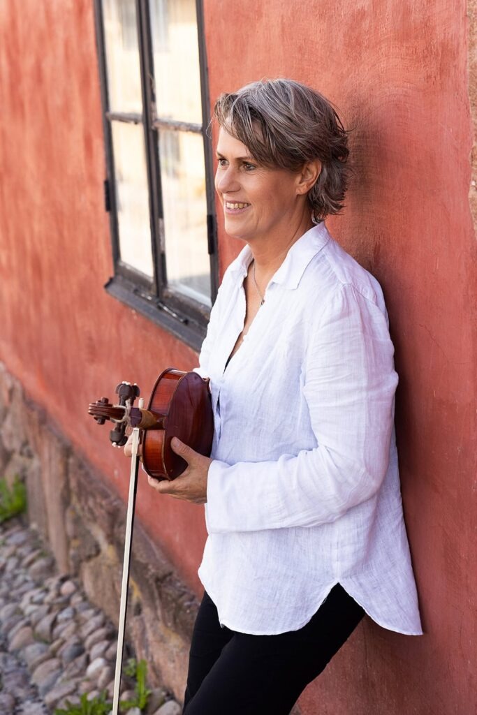 Leende kvinna i vit skjorta håller i en fiol och lutar sig mot en röd vägg nära ett fönster.