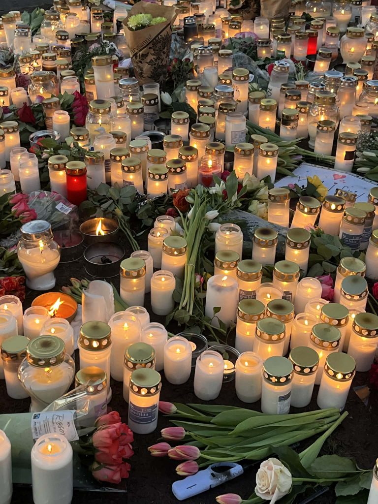 Många tända ljus och blommor placerade på marken i en minnesplats eller vaka, sett i skymningen.