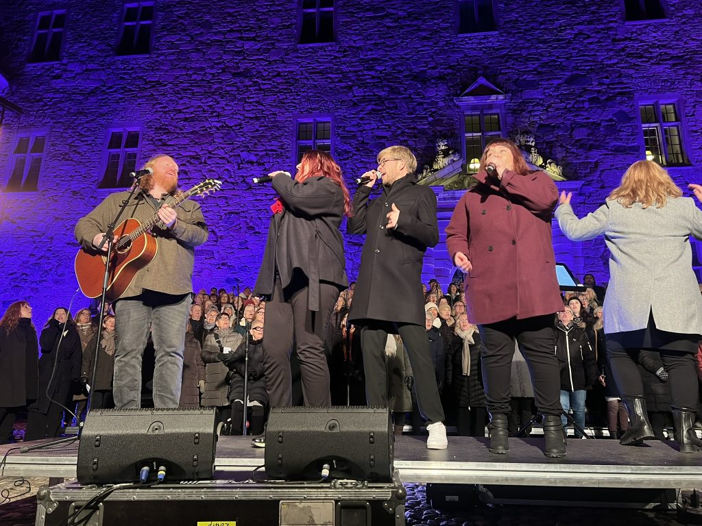 Fem personer sjunger och spelar gitarr på scenen på natten med en kör och en stenbyggnad upplyst i lila bakom dem.