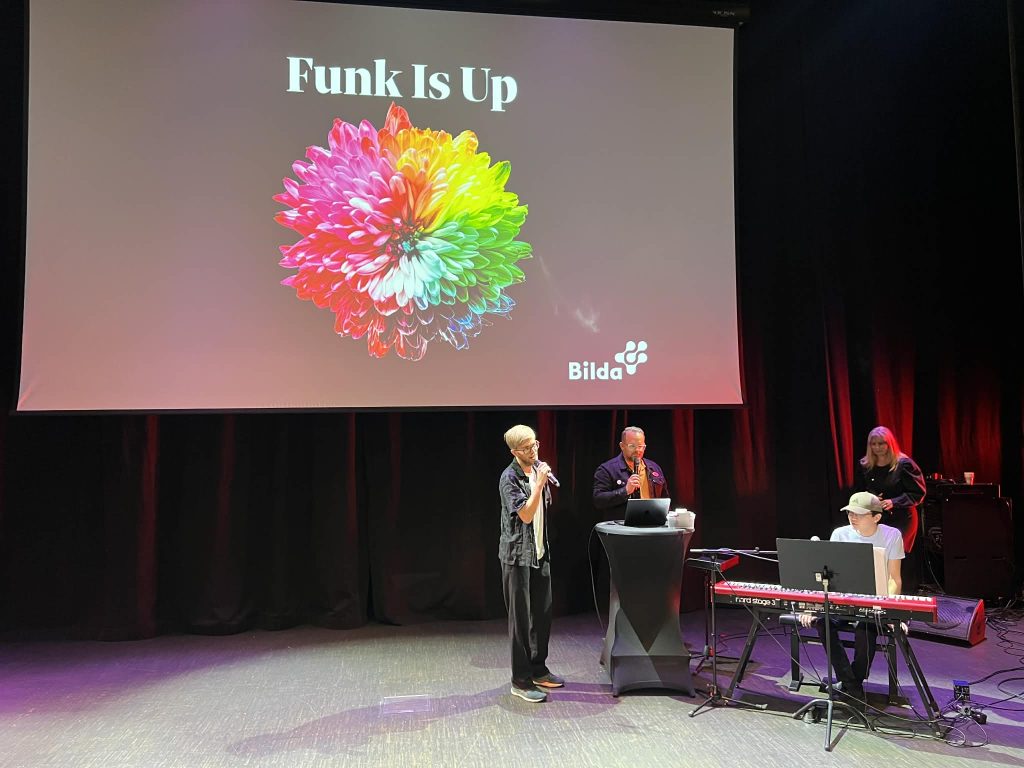 Fyra personer uppträder på scenen under en skärm som visar "Funk Is Up" och en färgglad blomstergrafik.