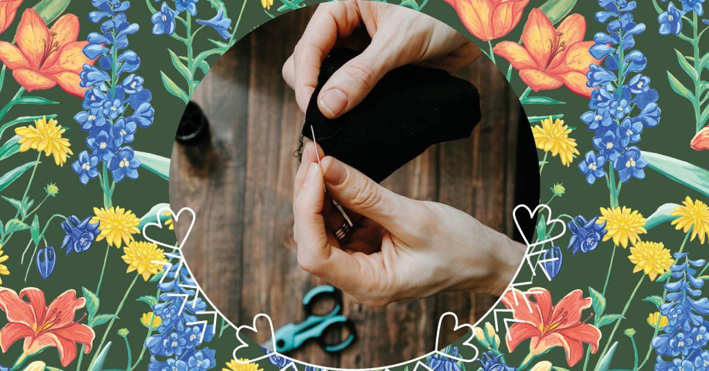 Händer som syr svart tyg med en nål, omgiven av färgglada blommor och hjärtbård, sax i närheten.