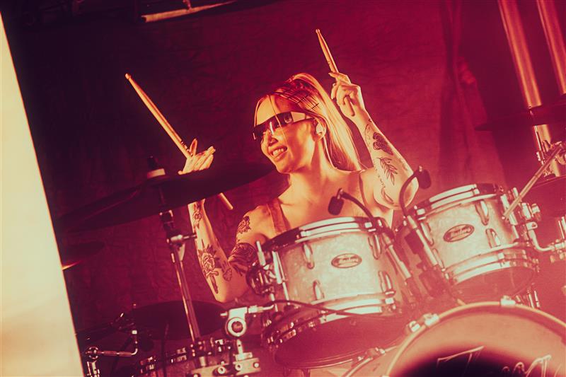 En leende kvinna med tatueringar spelar trummor på scenen, håller i trumpinnar och bär solglasögon.