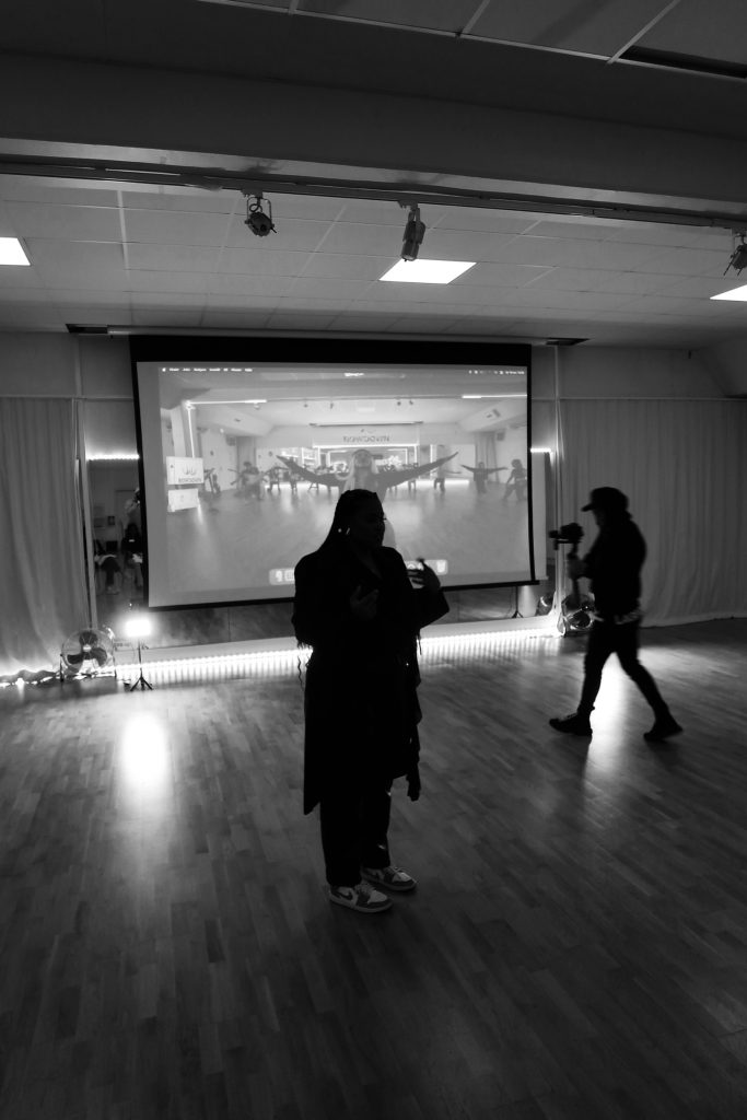 Två personer står framför en stor skärm som visar en video i ett mörkt, rymligt rum.