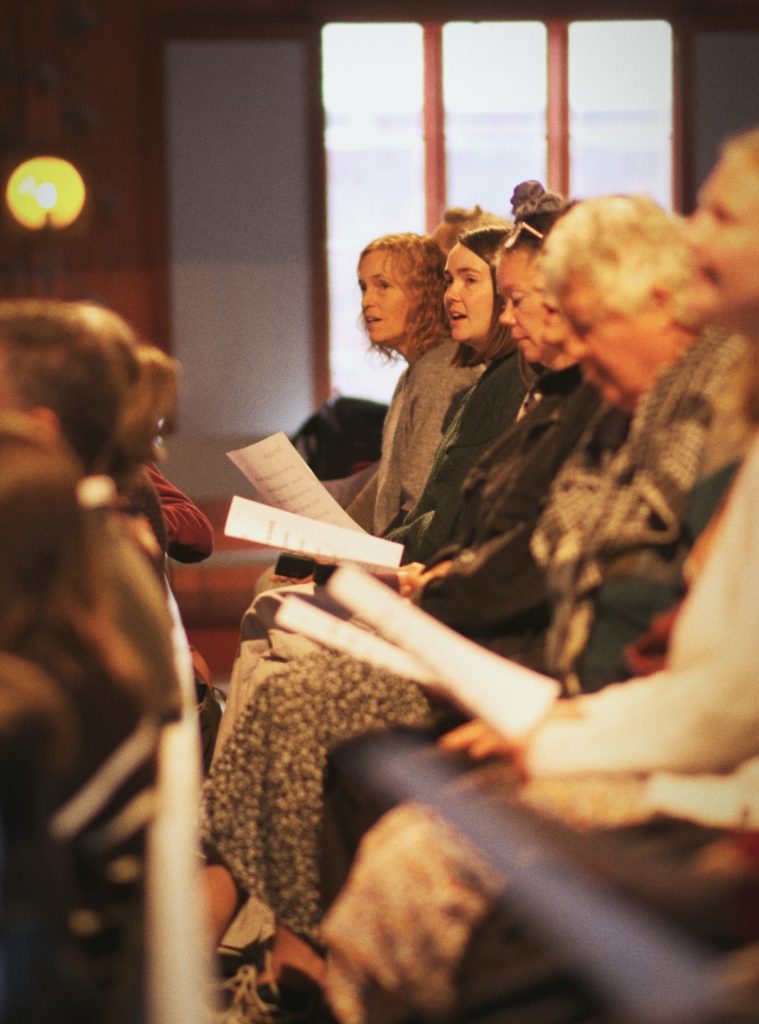 Människor sitter i rader inomhus, håller i papper, sjunger kanske eller deltar i ett framträdande eller en gudstjänst.