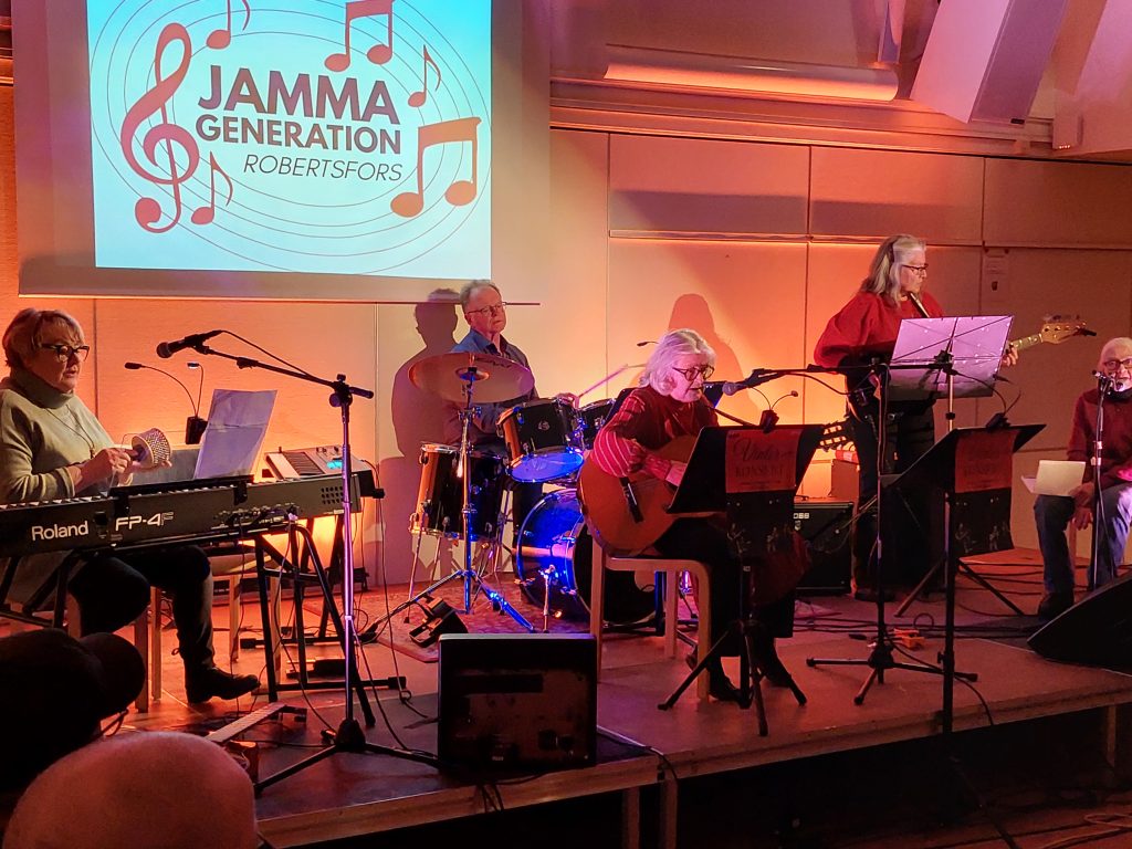 Ett band med äldre vuxna som uppträder på scen med instrument under en skylt med texten "Jamma Generation Robertsfors.