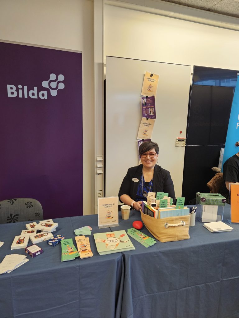 En person sitter i ett infobås med broschyrer och en korg framför en lila "Bilda"-banderoll.