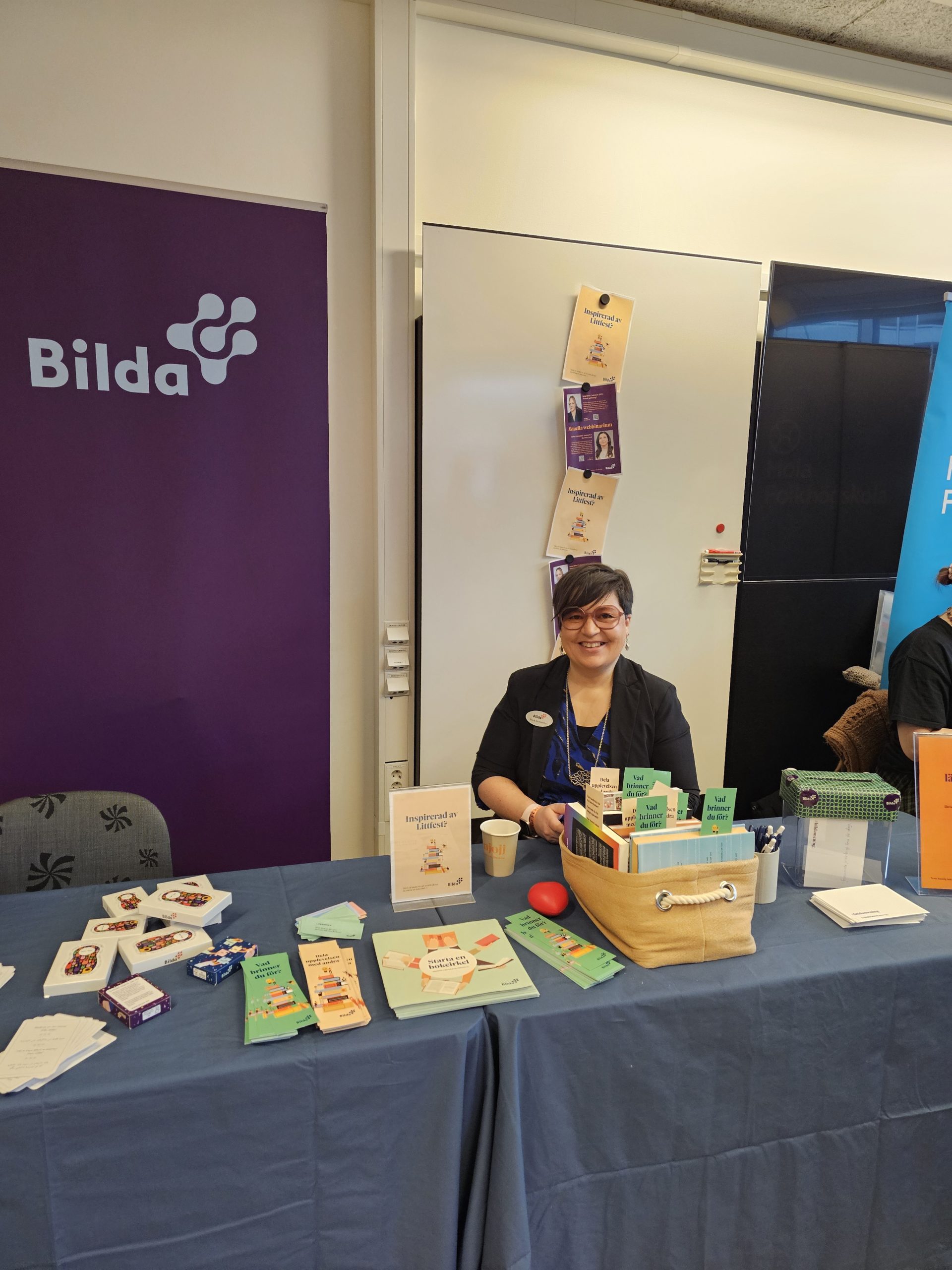 En person sitter i ett infobås med broschyrer och en korg framför en lila "Bilda"-banderoll.