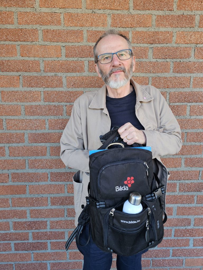 En äldre man med glasögon håller upp en svart Blida-ryggsäck mot en tegelvägg.