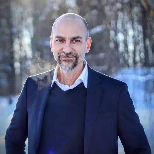 Skallig man med skägg i kostym, stående utomhus på vintern med träd och snö i bakgrunden.