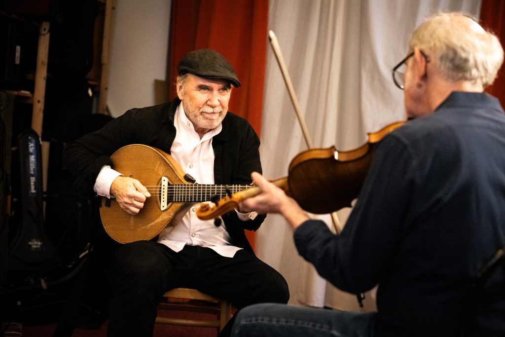 Två äldre män spelar stränginstrument tillsammans; den ene spelar mandolin, den andre spelar violin inomhus. Foto: Mats Lefvert