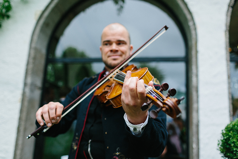 En man i traditionell klädsel spelar violin utomhus framför ett välvt fönster.