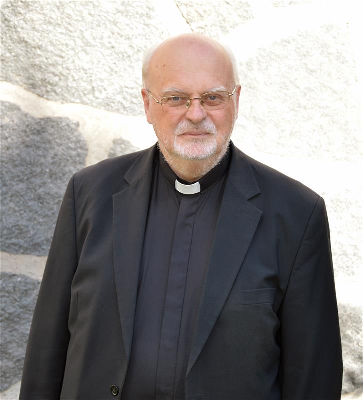 En äldre man med glasögon och prästkrage står framför en stenmur.